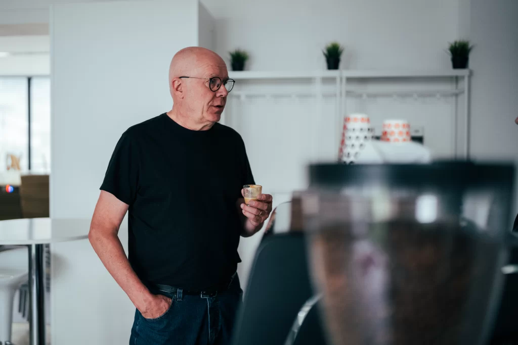 Muž v černém tričku drží šálek při setkání v kanceláři.