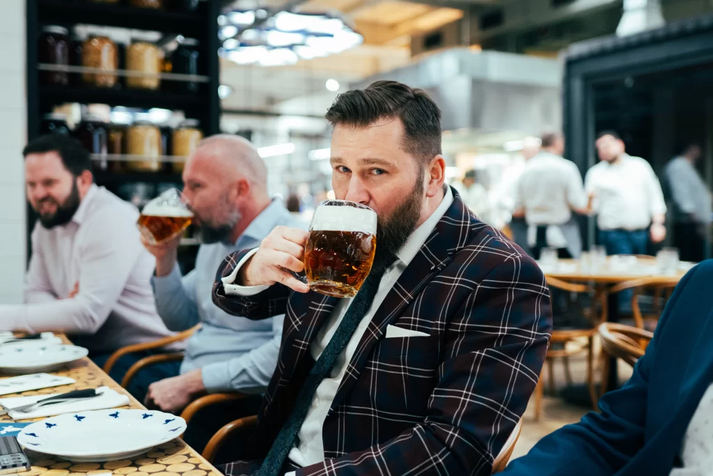 Muž v kostkovaném saku pije pivo u společného stolu.