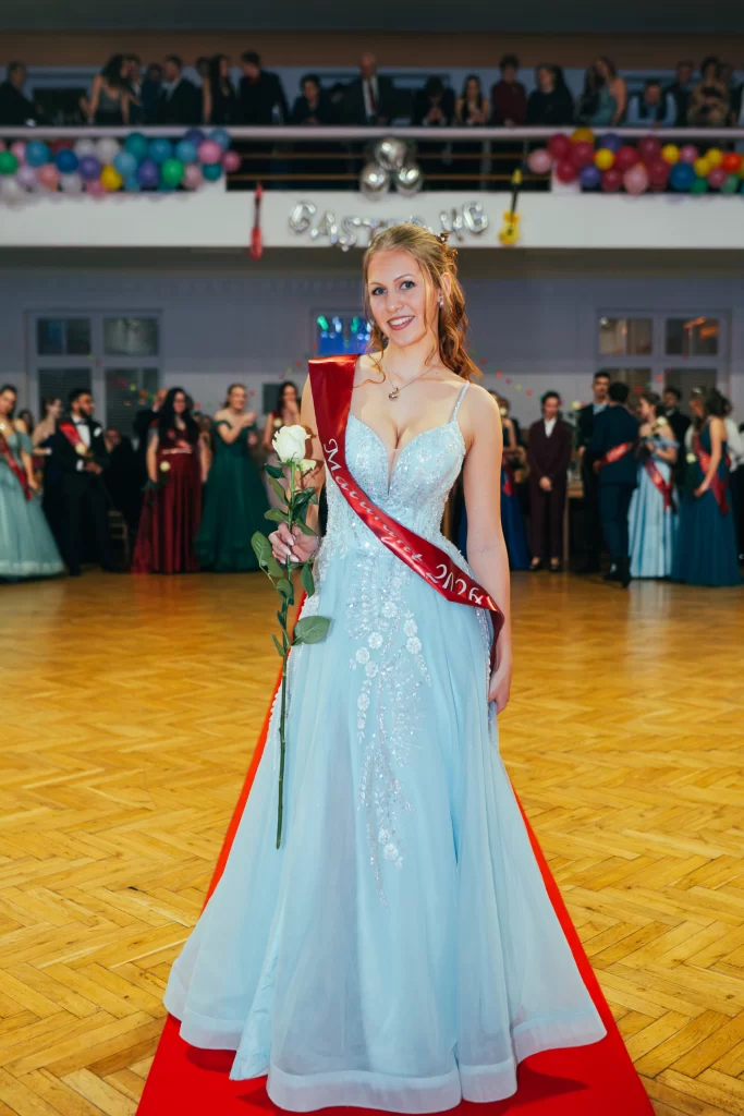 Maturantka ve světle modrých šatech s červenou šerpou drží růži.