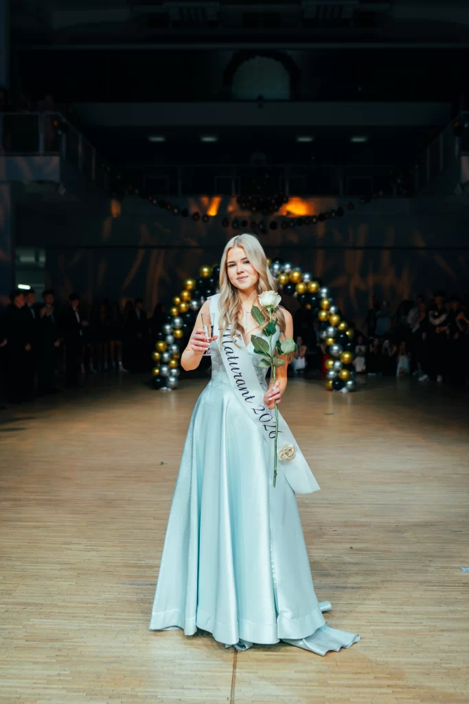 Maturantka s bílou šerpou drží sklenici a růži před balónkovým obloukem.